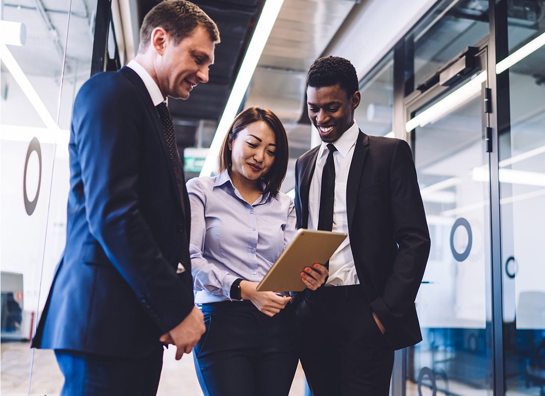 Contact - Portrait of a Group of Three Smiling Business Colleagues Standing Together in a Hallway in the Office While Looking at a Tablet